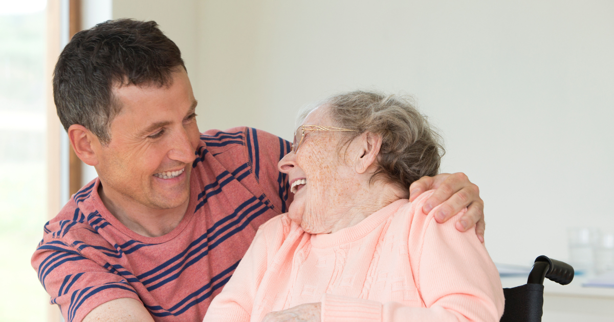 Young man has his arm over the shoulder of an elderly woman in a wheelchair as she laughs