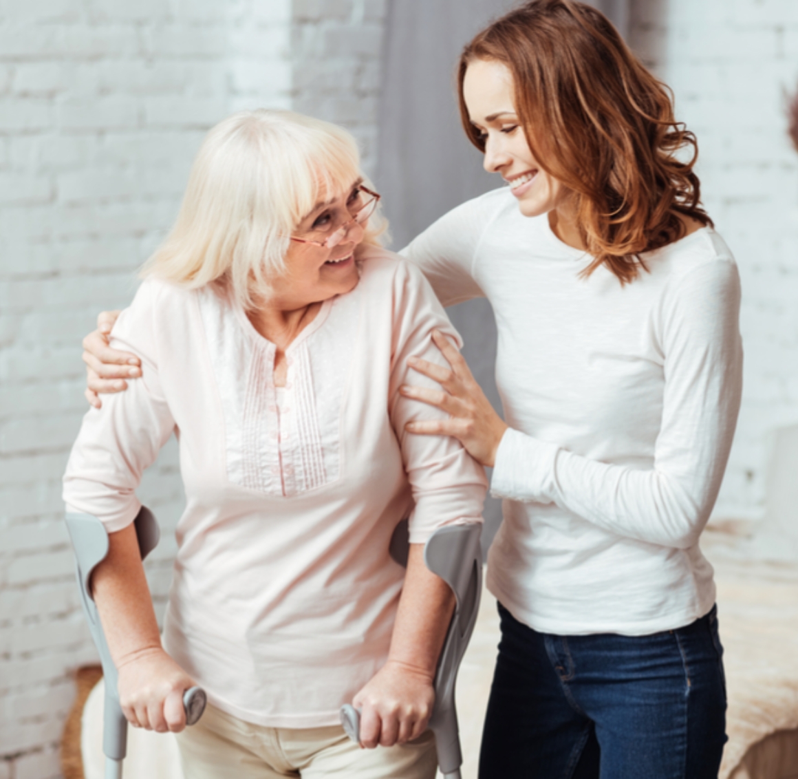 A smiling young woman supporting an elderly woman using forearm crutches. The elderly woman, with white hair and glasses, is dressed in a light pink top, while the younger woman, wearing a white long-sleeve shirt and jeans, gently holds her arm, both sharing a warm, encouraging moment indoors.