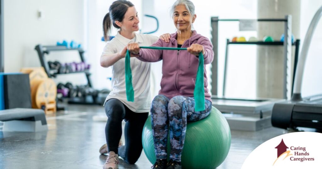 A care provider helps an older woman exercise with a resistance band and an exercise ball, representing how exercise can help with senior fall prevention.